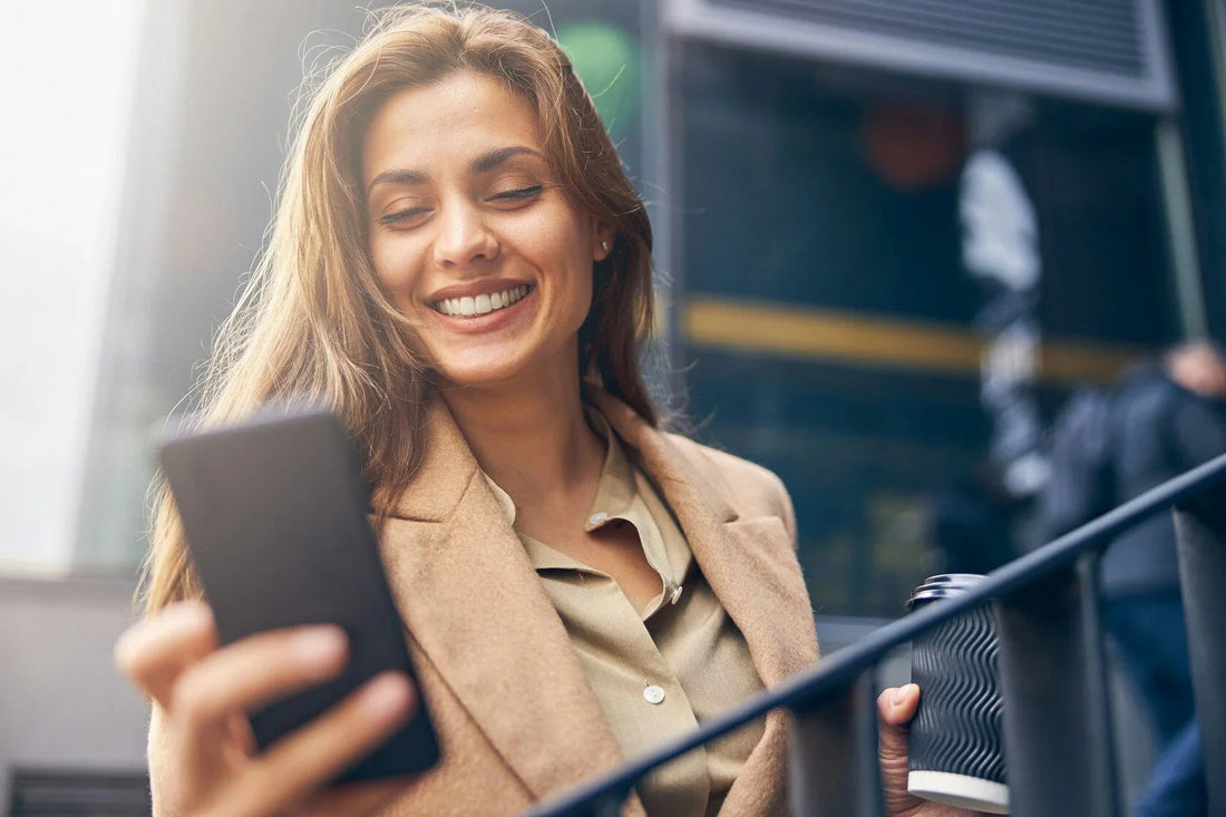 Vrouw in beige jas lacht terwijl ze een telefoon vasthoudt en een koffie beker buiten drinkt