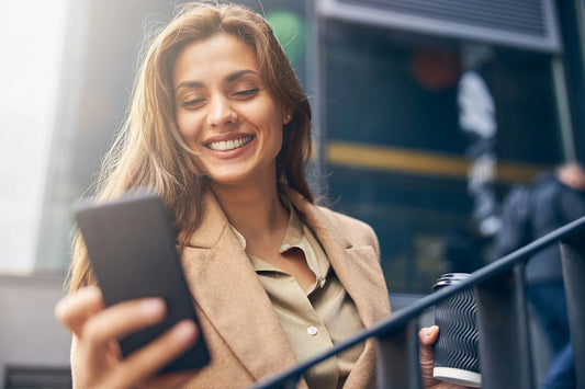 Vrouw in beige jas lacht terwijl ze een telefoon vasthoudt en een koffie beker buiten drinkt