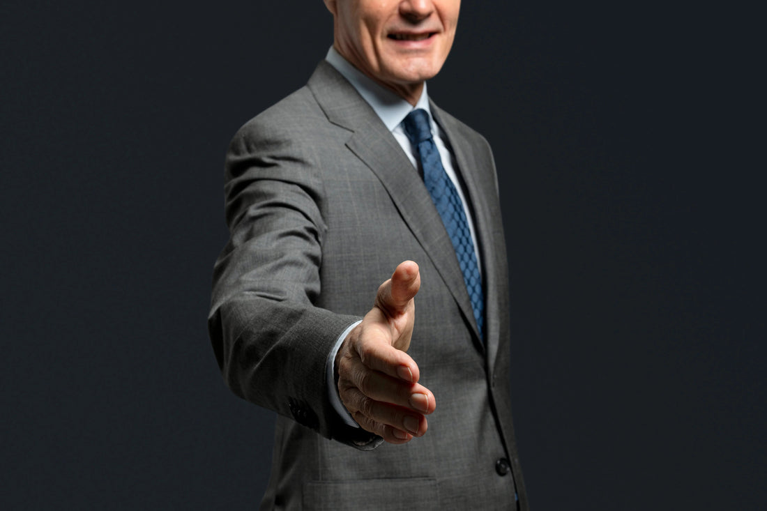 Zakelijke man in grijs pak met blauwe stropdas die hand uitsteekt voor een handdruk, donkere achtergrond